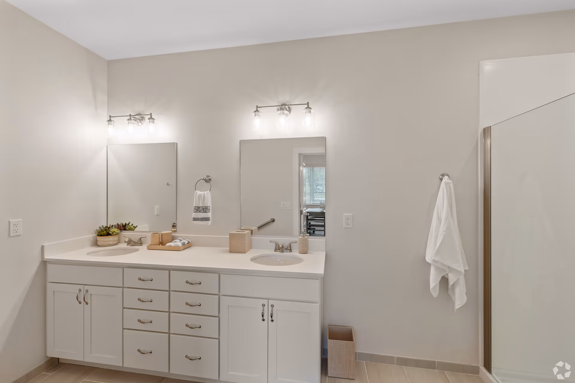 A clean and modern bathroom featuring a double sink vanity with white cabinets and drawers. Above each sink is a rectangular mirror with light fixtures mounted above them. A small potted plant and neatly arranged toiletries are placed on the countertop. A white towel hangs on a ring beside the left mirror, and another white towel hangs on a hook near a glass-enclosed shower on the right side of the image. The walls are painted light beige, and the floor has light-colored tiles.