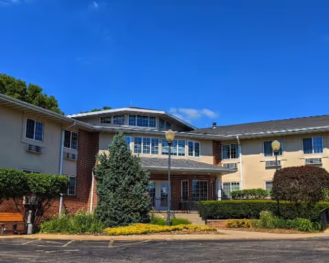 Exterior view of a two-story assisted living facility building with beige and brick walls, multiple windows, and a main entrance with a covered porch. There are shrubs, trees, and landscaped bushes in front of the building under a clear blue sky.