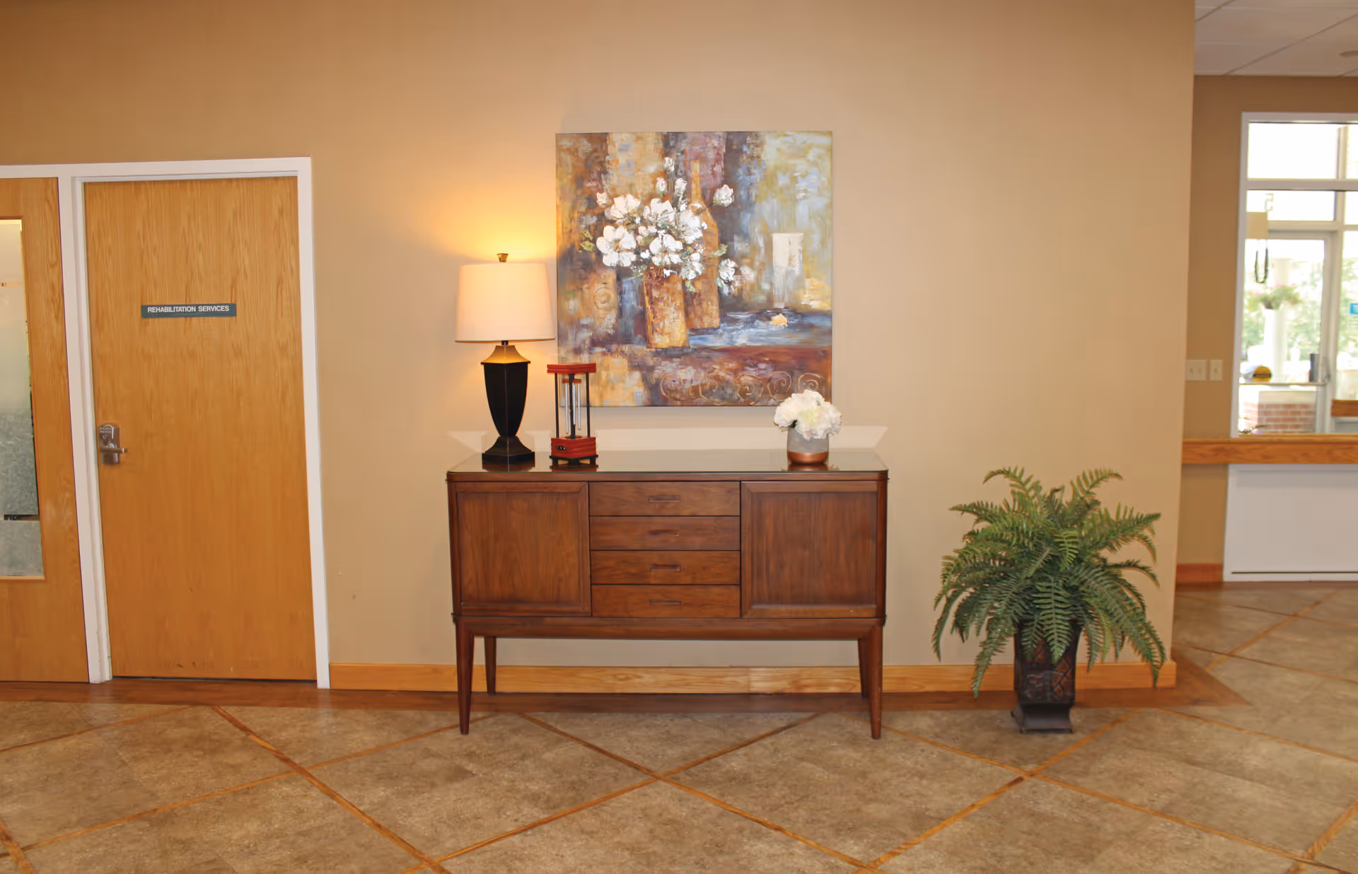 Interior view of a hallway in a senior living facility with a wooden sideboard against a beige wall. On the sideboard are a table lamp, a small decorative red lantern, and a white flower arrangement. Above the sideboard hangs a painting of flowers. To the right of the sideboard is a potted fern plant. On the left side, there is a wooden door labeled 'Rehabilitation Services'. The floor is tiled with a brown and beige pattern.