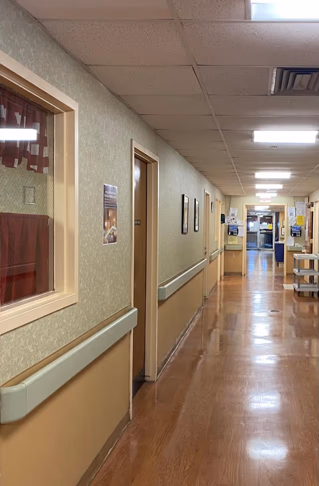 A long, clean hallway inside a senior living facility with beige walls, wooden doors, handrails along the walls, and a polished wooden floor. The hallway is well-lit with ceiling lights and has various notices and posters on the walls. At the end of the hallway, there is a glass door leading to another area.