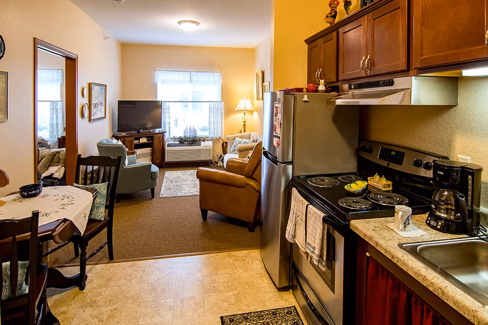 View of a cozy living space in a senior living facility showing a small kitchen area with a stove, refrigerator, and sink on the right, and a dining table with chairs on the left. Beyond the kitchen is a living room with armchairs, a TV on a stand, a window with curtains, and a lamp providing warm lighting.