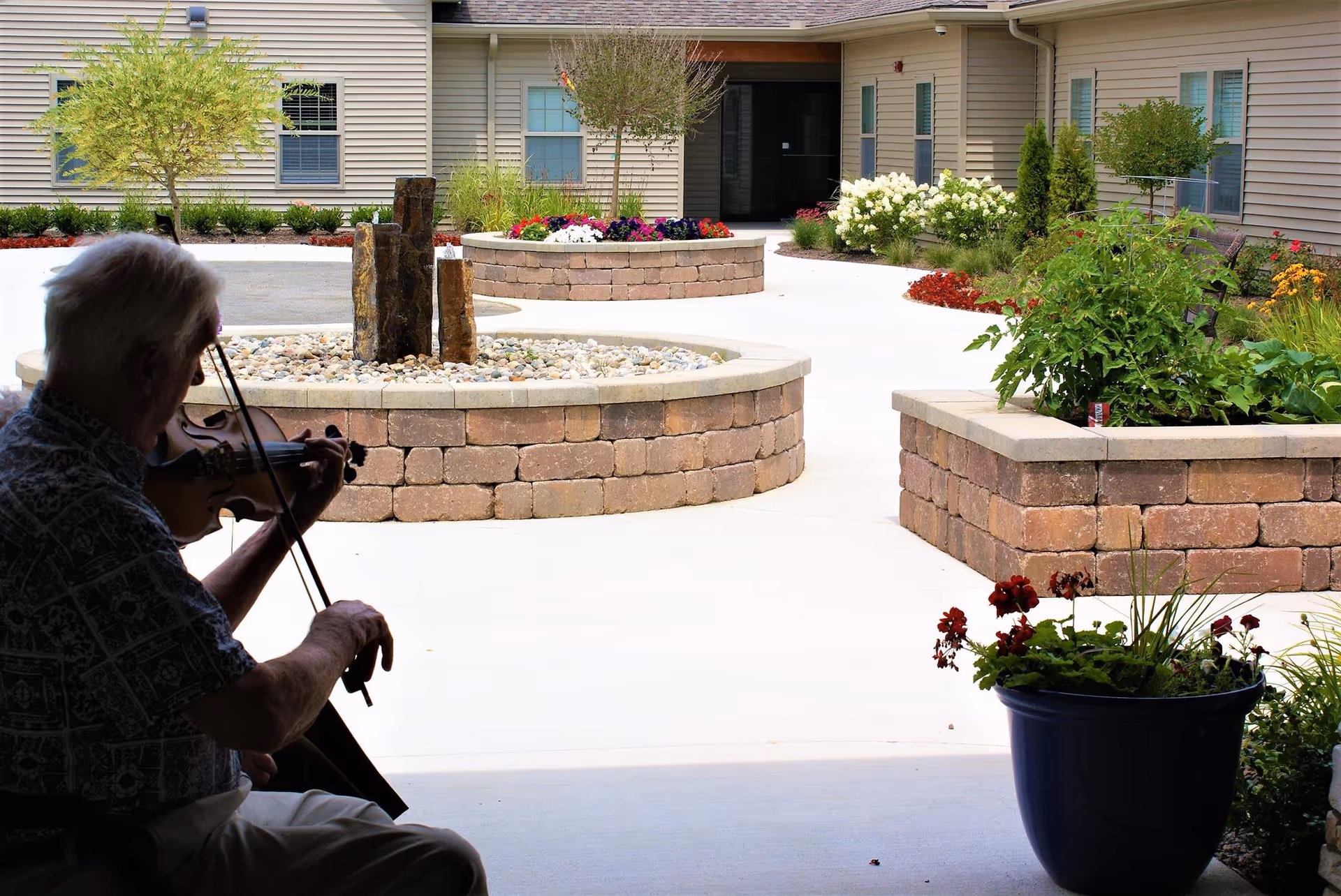 An elderly person playing a violin in a courtyard garden area of an assisted living facility. The courtyard features raised stone planters with flowers and small trees, surrounded by a beige building with multiple windows.