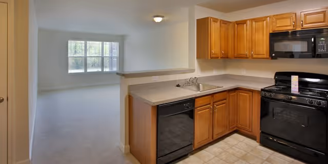 Interior view of a senior living facility apartment showing a kitchen area with wooden cabinets, a black stove, microwave, dishwasher, and a sink. The kitchen opens into a bright living room with large windows letting in natural light.