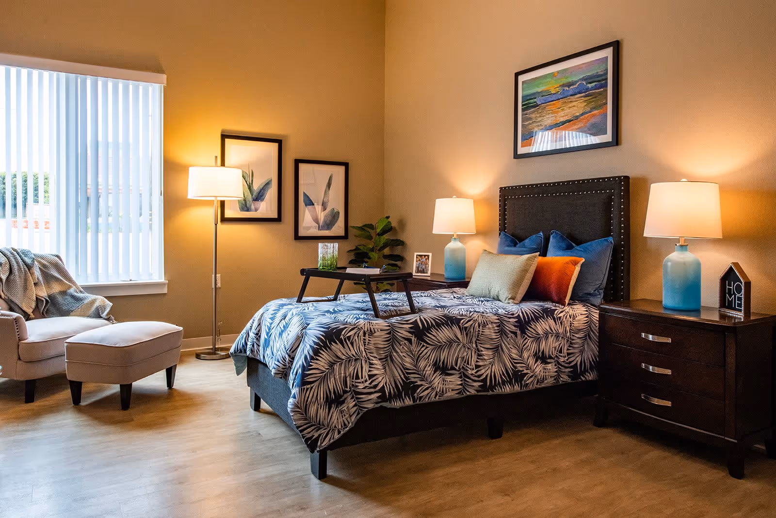 A cozy bedroom with a bed featuring a dark upholstered headboard and a blue and white leaf-patterned bedspread. The bed has multiple pillows in blue, orange, and beige colors. There are two dark wooden nightstands on either side of the bed, each with a blue lamp and decorative items. On the left side of the room, there is a beige armchair with a matching ottoman near a window with vertical blinds. The walls are painted a warm beige color and decorated with framed artwork. The floor is light wood.