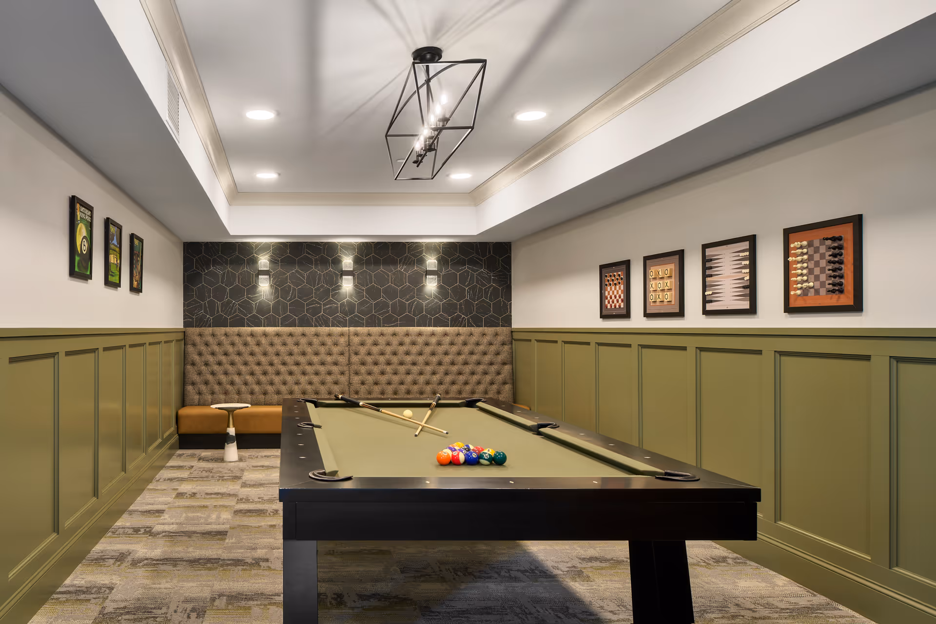 A recreational room with a pool table in the center, two pool cues resting on the table, and a set of billiard balls arranged for a game. The room has green wainscoting on the walls, a tufted bench along the back wall, and framed game-themed artwork including chess and backgammon boards. The ceiling features recessed lighting and a modern rectangular chandelier.