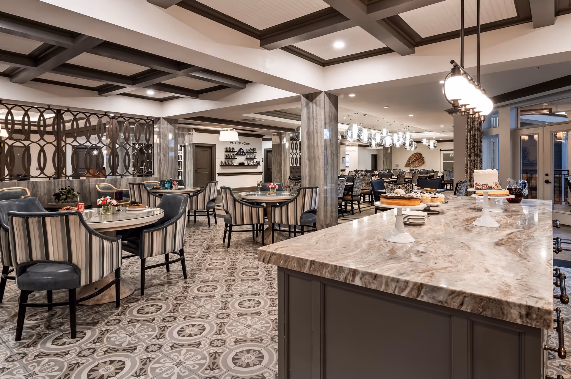 A spacious and elegant dining area in The Remington of McCandless featuring round tables with striped and leather chairs, a large marble countertop island with cakes and pastries displayed, decorative ceiling beams, patterned tile flooring, and modern pendant lighting. The room has a warm and inviting atmosphere with a 'Wall of Honor' visible in the background.