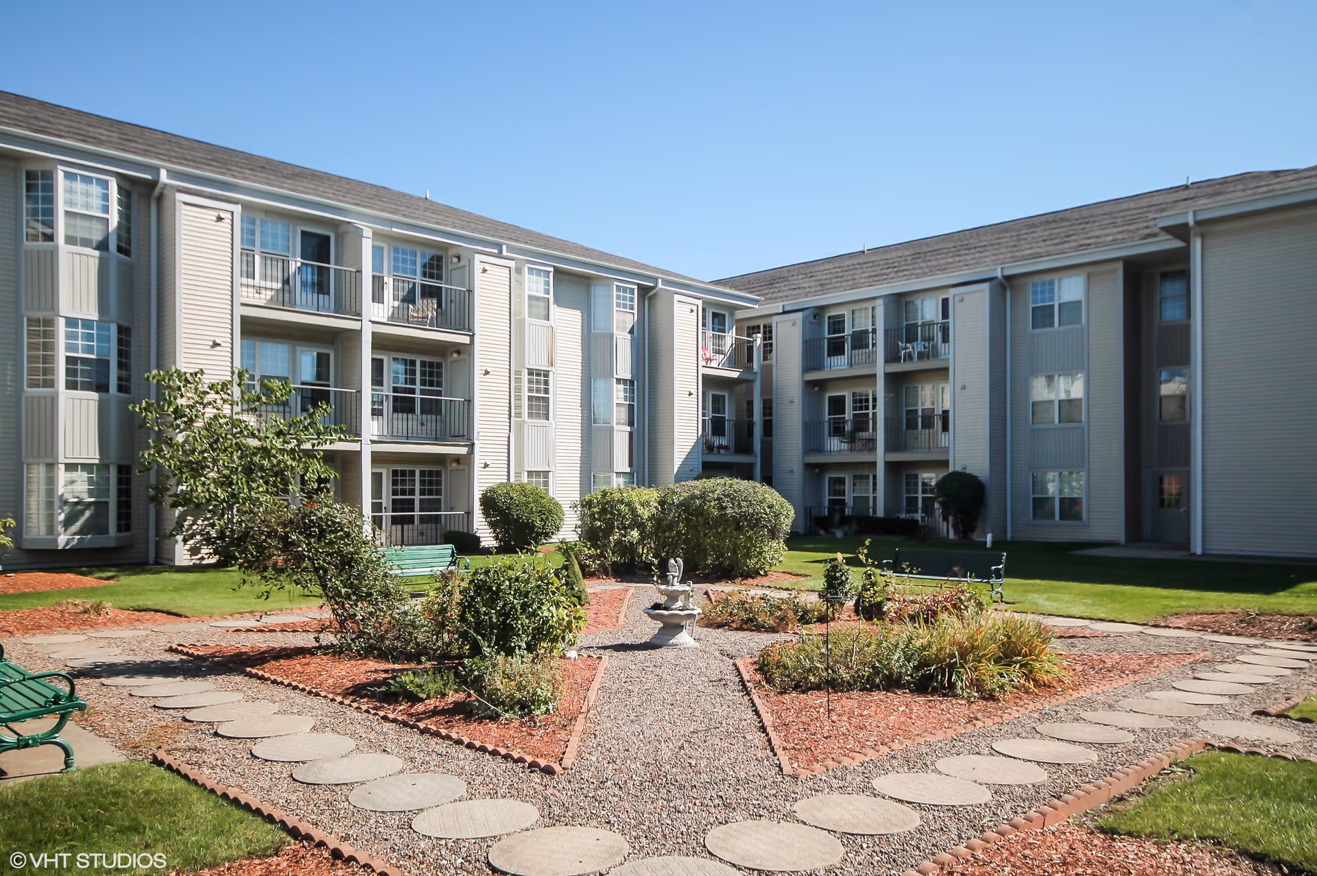 Outdoor courtyard area of a senior living facility with a landscaped garden featuring a small fountain in the center, surrounded by bushes and plants. The courtyard is bordered by a three-story building with balconies and windows. There are green benches placed around the garden area under a clear blue sky.