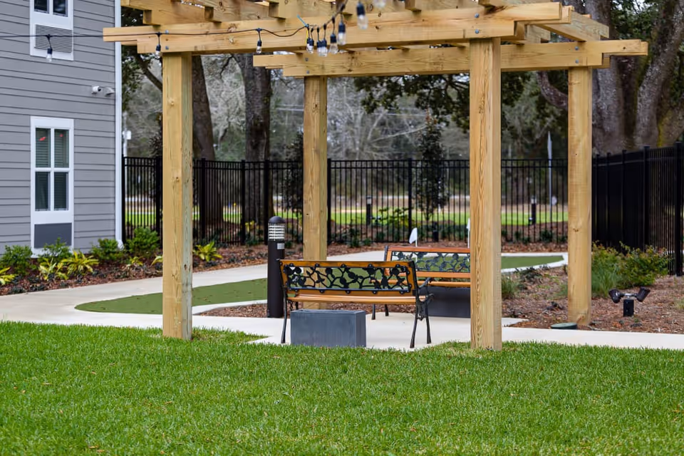 Outdoor seating area with two wooden benches under a wooden pergola structure with string lights, surrounded by green grass, a paved walkway, and landscaping with trees and shrubs in the background.