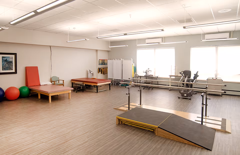A spacious physical therapy room with wooden flooring, two padded therapy tables, exercise balls, parallel bars with a ramp, and exercise equipment near large windows letting in natural light.