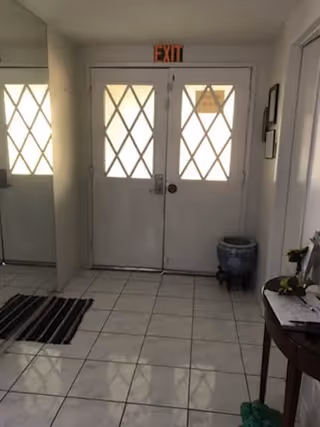Interior view of an entryway with double doors featuring diamond-patterned windows. There is an exit sign above the doors, a small round decorative pot on the floor to the right, a striped rug on the tiled floor to the left, and a small table with papers and a plant on the right side.