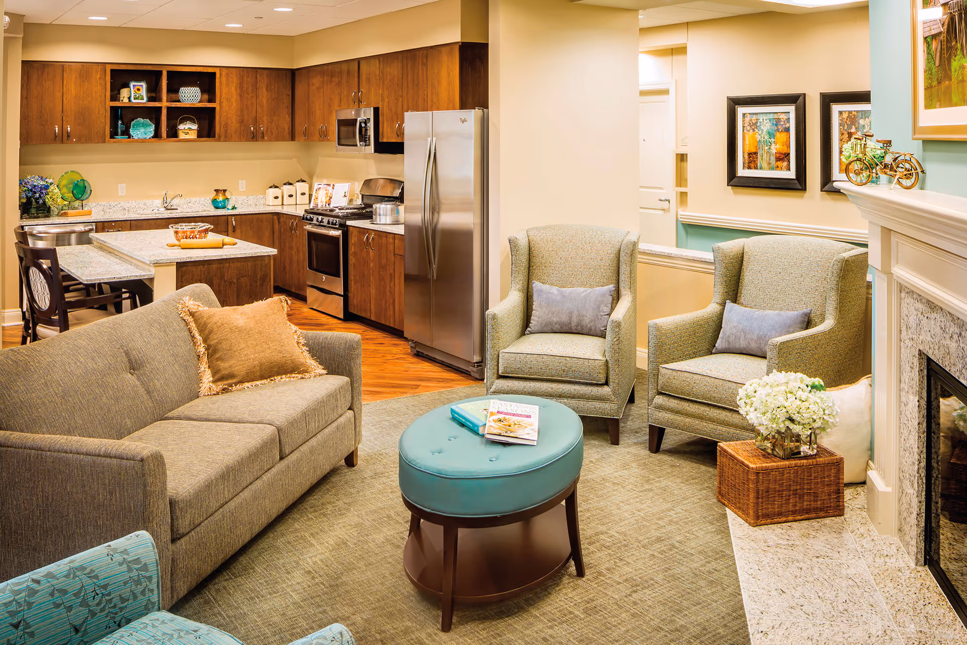 A cozy living room and kitchen area featuring a gray sofa with a brown pillow, two green armchairs with gray cushions, a round teal ottoman with books on top, and a fireplace with decorative items. The kitchen has wooden cabinets, a stainless steel refrigerator, oven, microwave, and a small island with two chairs.
