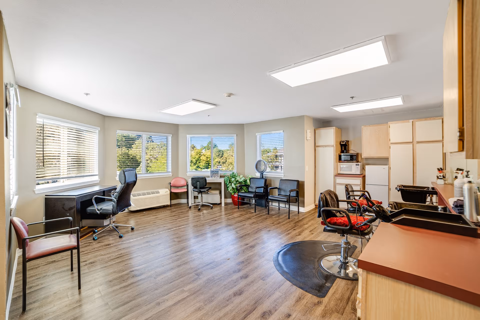 Bright multipurpose interior room with large windows, several chairs, salon styling stations and a small kitchenette area.