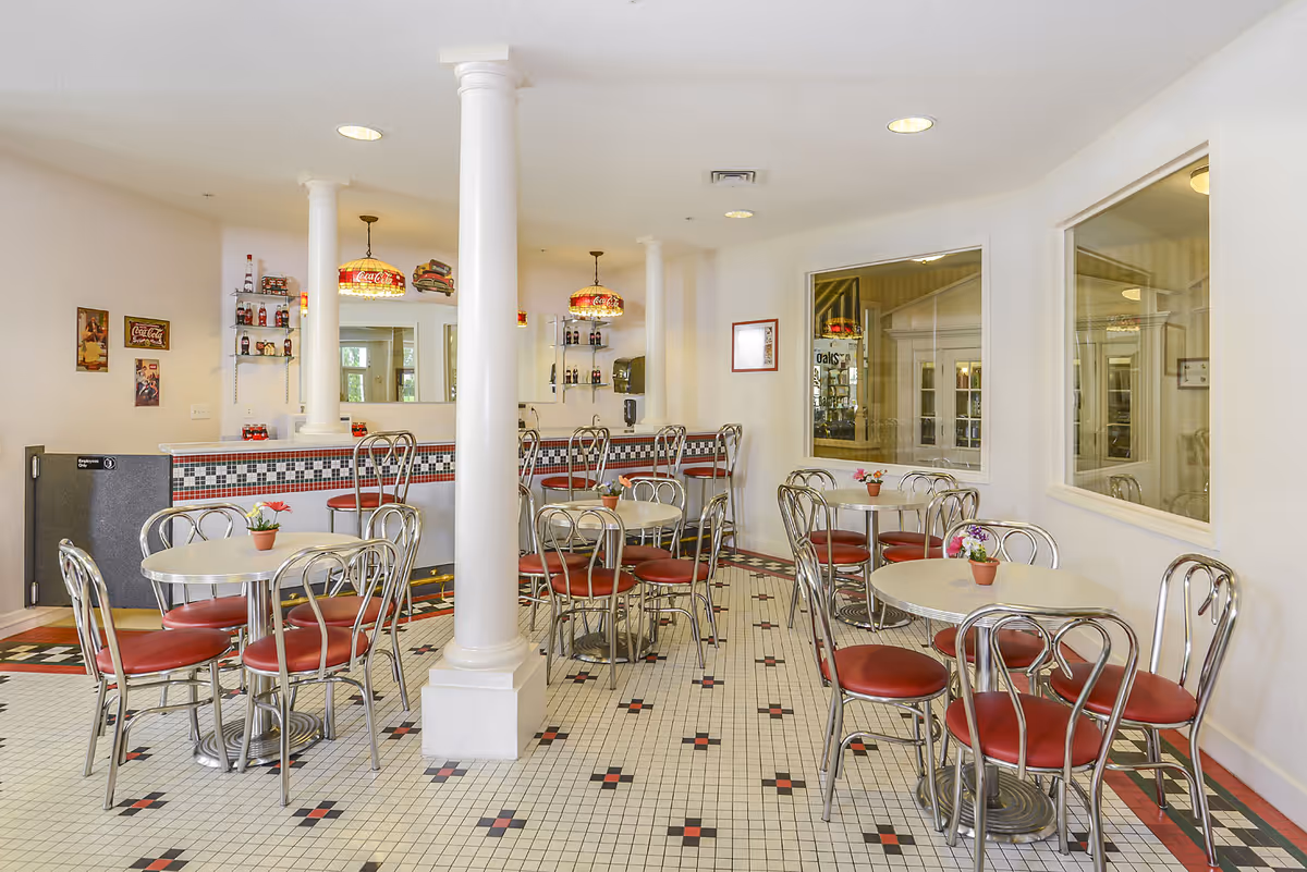 Bright retro-style dining area with round tables, red-cushioned chrome chairs, tiled floor, and a counter with pendant lights.