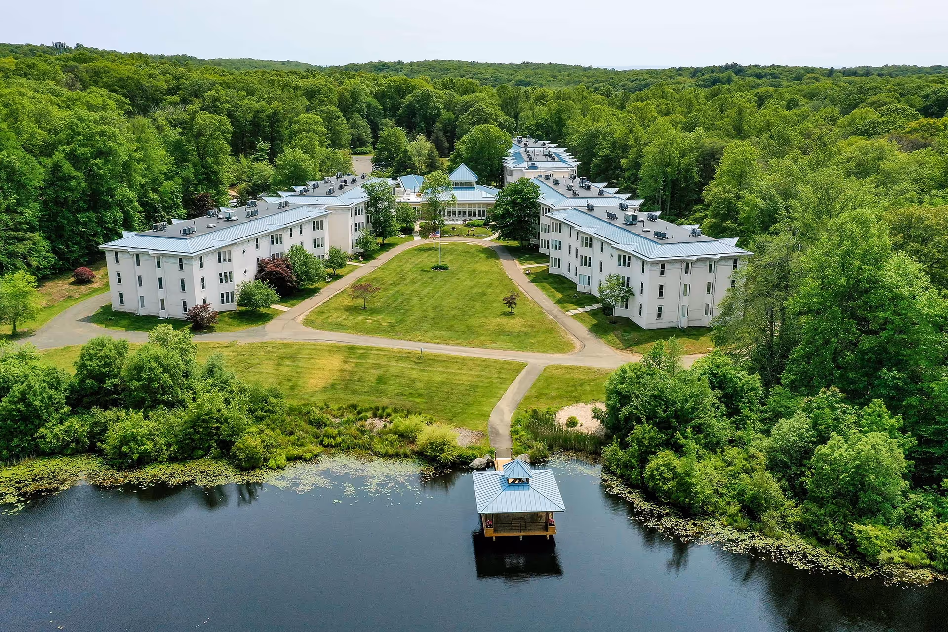 Aerial view of Guilford Retirement Residence showing two long, white multi-story buildings with blue roofs facing each other across a large green lawn with a flagpole in the center. The buildings are surrounded by dense green trees and a small lake with a covered dock is visible in the foreground.