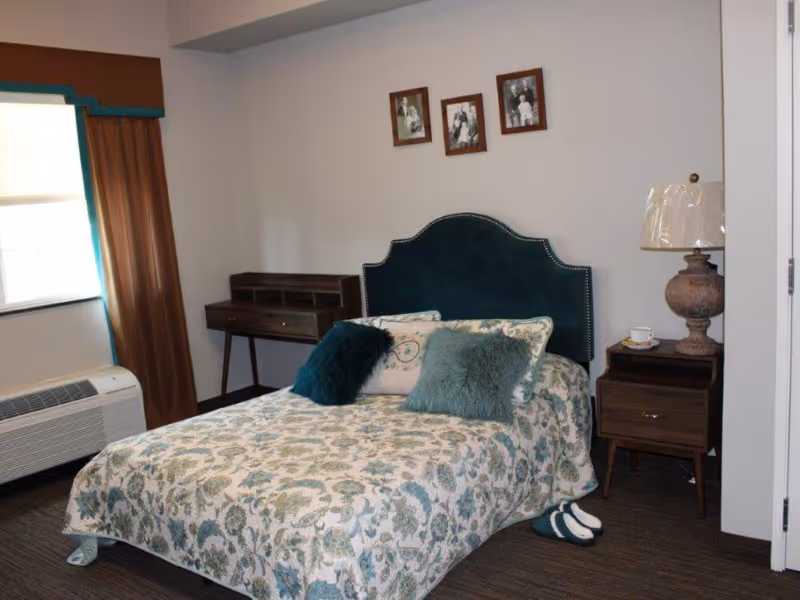 A bedroom with a bed covered in a floral patterned bedspread and three decorative pillows. There is a dark green upholstered headboard, a wooden nightstand with a lamp and a cup, a small wooden desk against the wall, and three framed black and white photos hanging above the bed. A window with brown curtains and an air conditioning unit is visible on the left side.