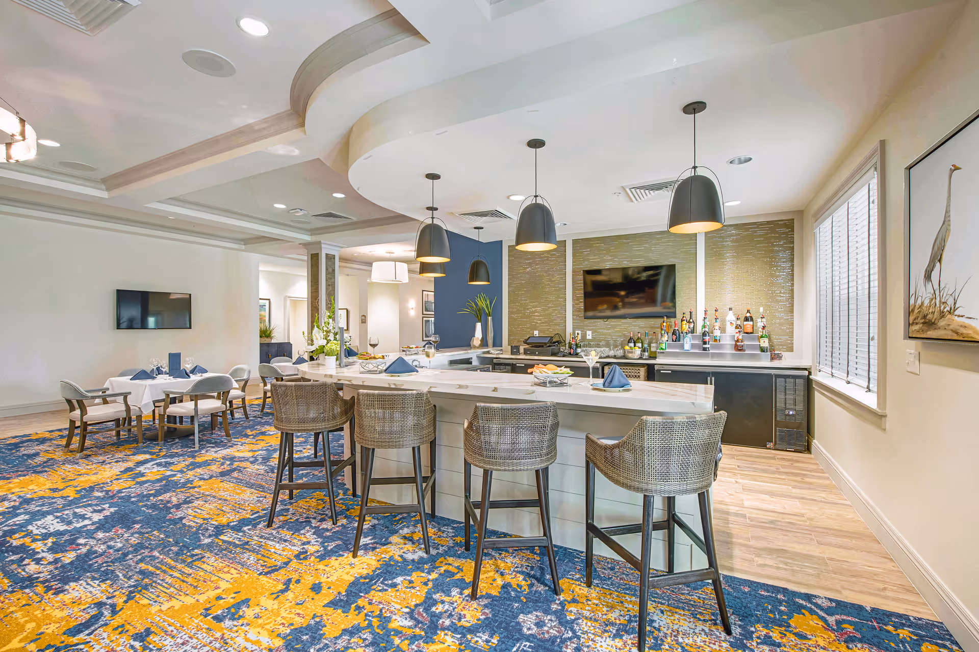 Spacious indoor dining and lounge area featuring a central bar island with stools, pendant lights, dining tables, and a colorful patterned carpet.