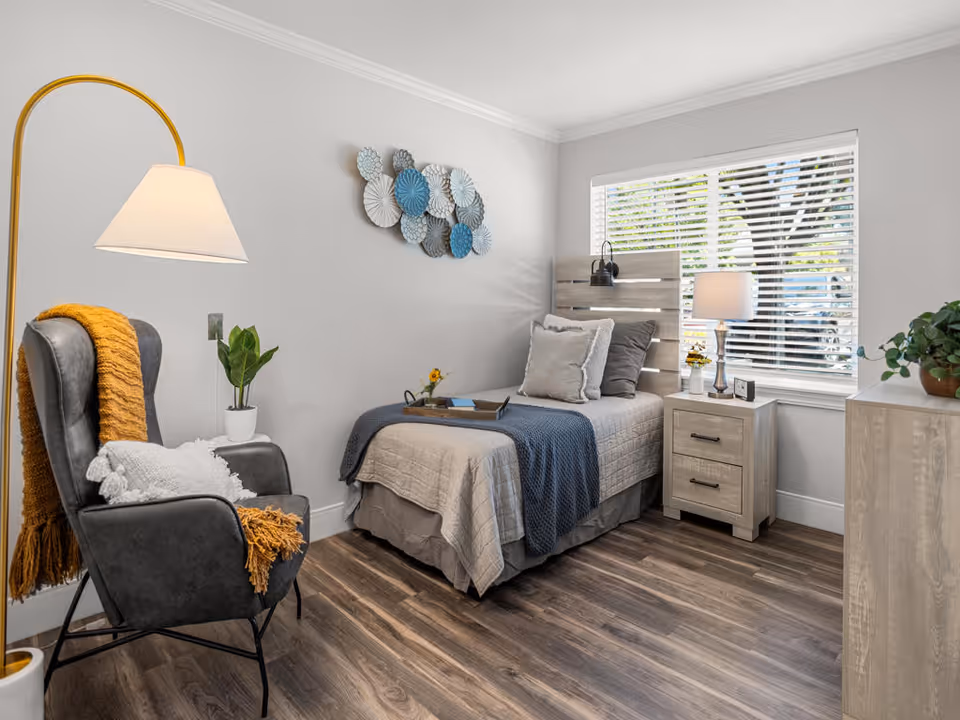 A cozy bedroom with a single bed dressed in beige and blue bedding, a wooden headboard, and two pillows. Next to the bed is a wooden nightstand with a lamp, a small plant, and a clock. A large window with white blinds lets in natural light. On the left side, there is a gray armchair with a white pillow and a mustard yellow throw blanket draped over it, accompanied by a tall floor lamp. The floor is wood, and the walls are painted light gray with decorative wall art above the bed.