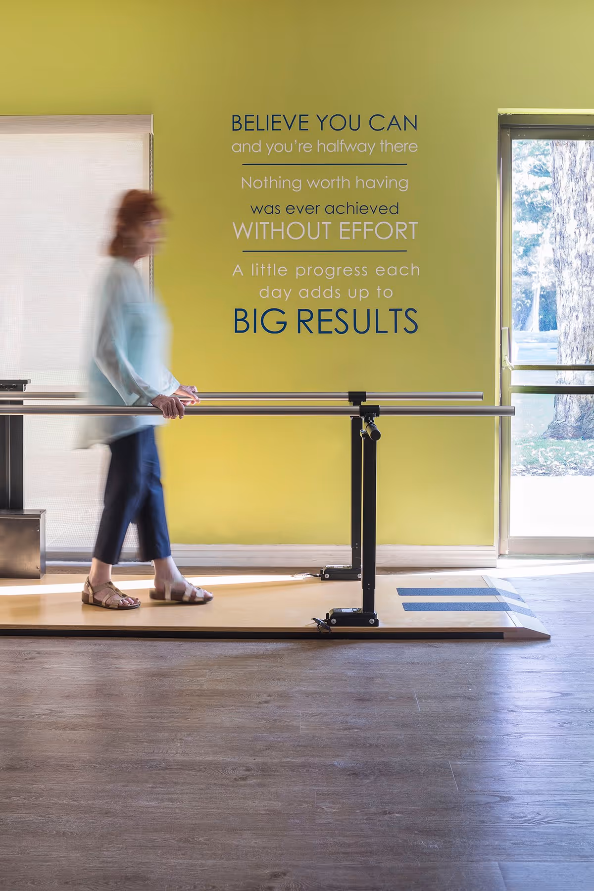 A senior woman walking with support on parallel bars in a rehabilitation or physical therapy room with a motivational quote on a yellow-green wall and a large window showing trees outside.