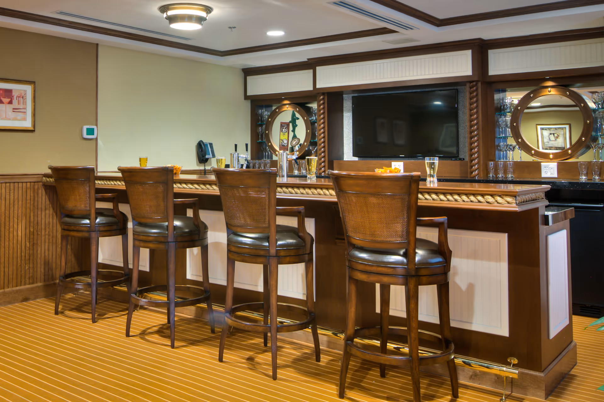 Interior view of a bar area with four wooden bar stools with leather seats lined up in front of a wooden bar counter. The bar has a rope detail along the edge and is set against a wall with two round mirrors and glass shelves holding glassware. A flat-screen TV is mounted on the wall behind the bar. The room has warm lighting and a striped carpeted floor.