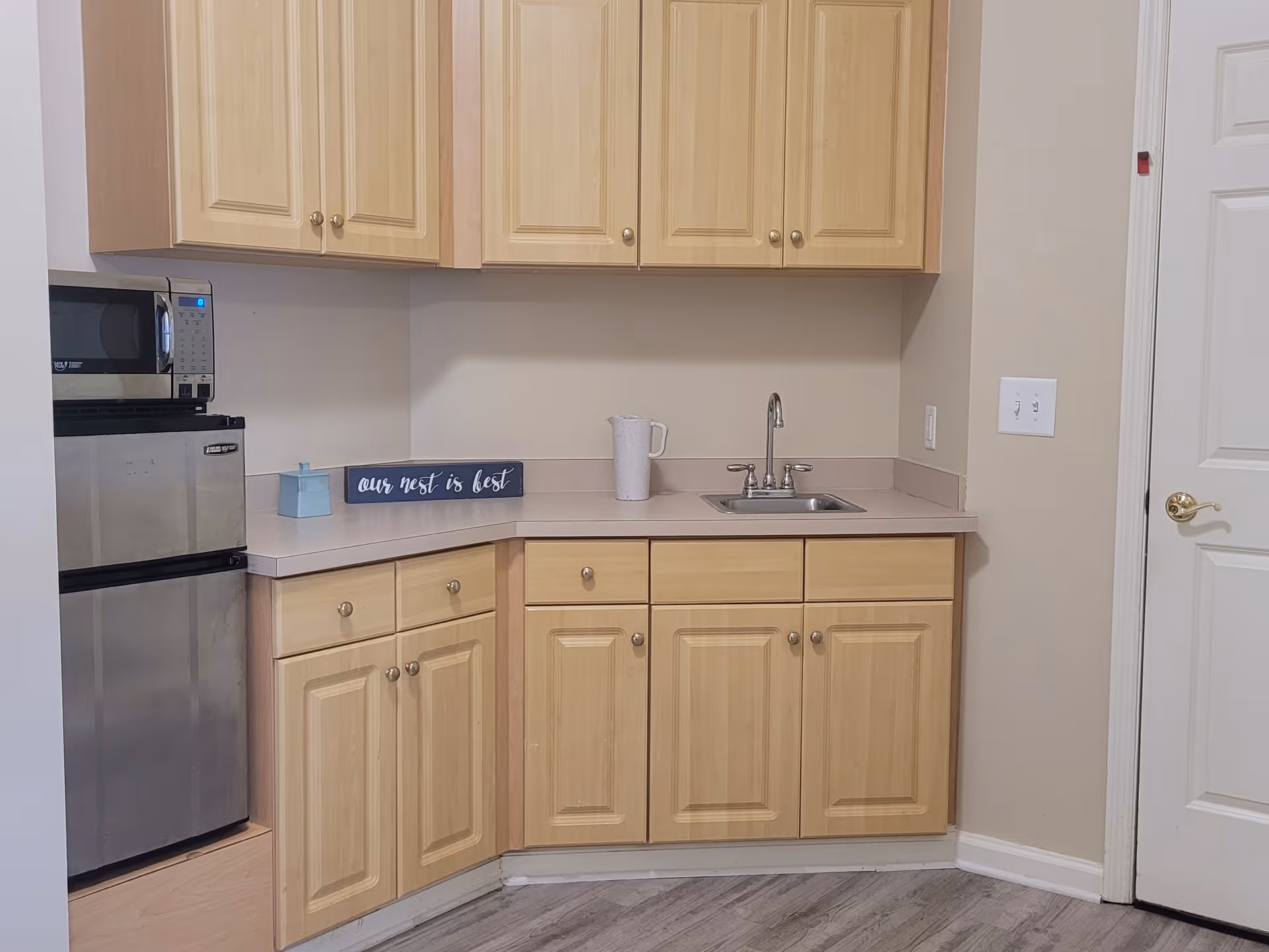 A small kitchen area with light wood cabinets, a stainless steel mini refrigerator, a microwave on top of the refrigerator, a countertop with a sink, a white pitcher, a small blue container, and a decorative sign that reads 'our nest is best'. The floor is light-colored wood, and there is a white door with a gold handle on the right side.