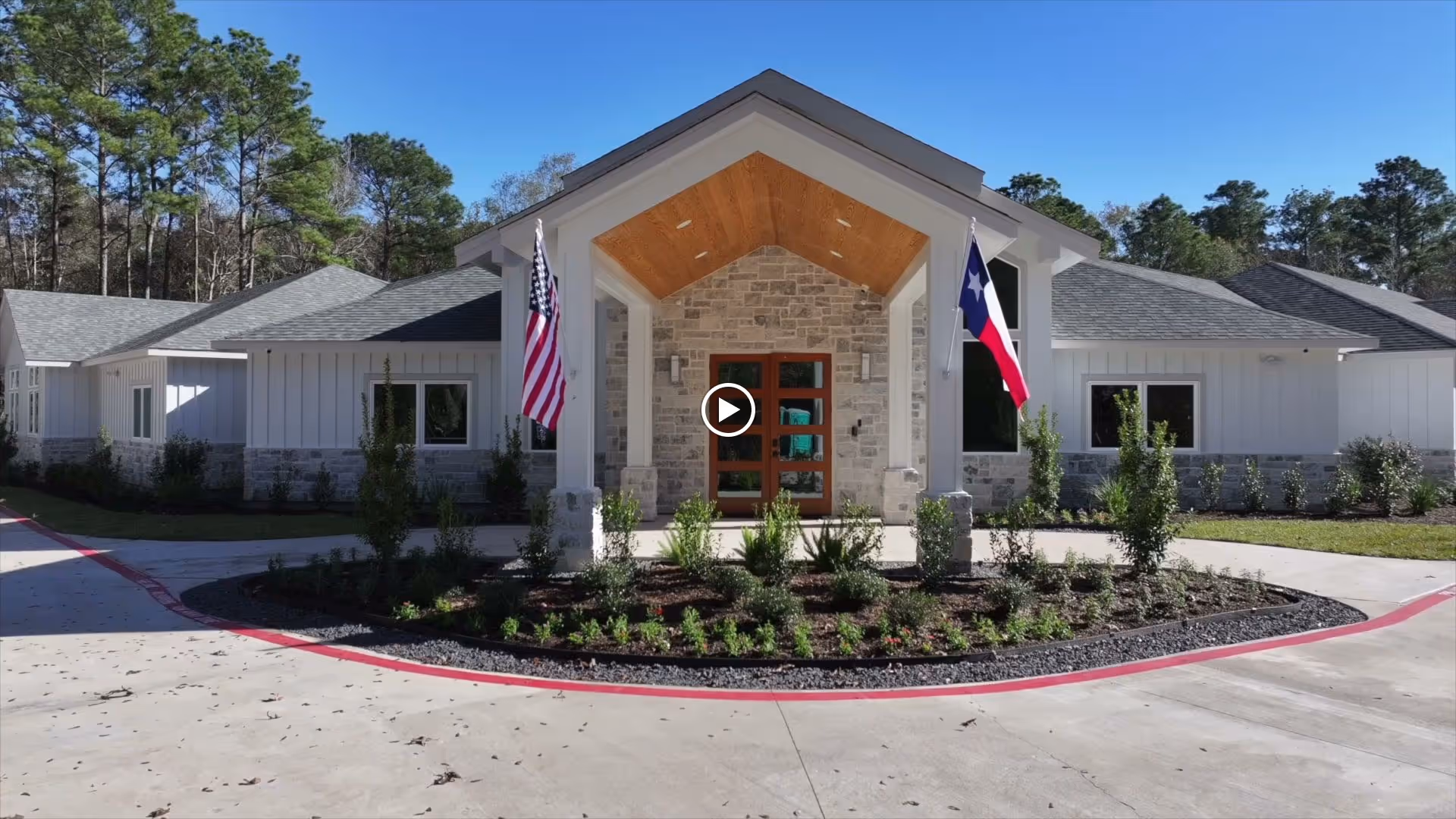 Front exterior view of The Magnolia Woods Assisted Living facility featuring a modern building with a peaked roof entrance, stone and white siding walls, two flagpoles displaying the American and Texas flags, and landscaped greenery in front.