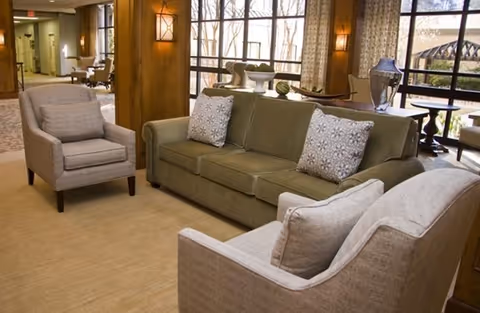 A cozy living room area with a green sofa and two beige armchairs arranged around a carpeted floor. The room features large windows with curtains, wooden pillars, and soft lighting from wall sconces. Outside the windows, trees and a patio area are visible.
