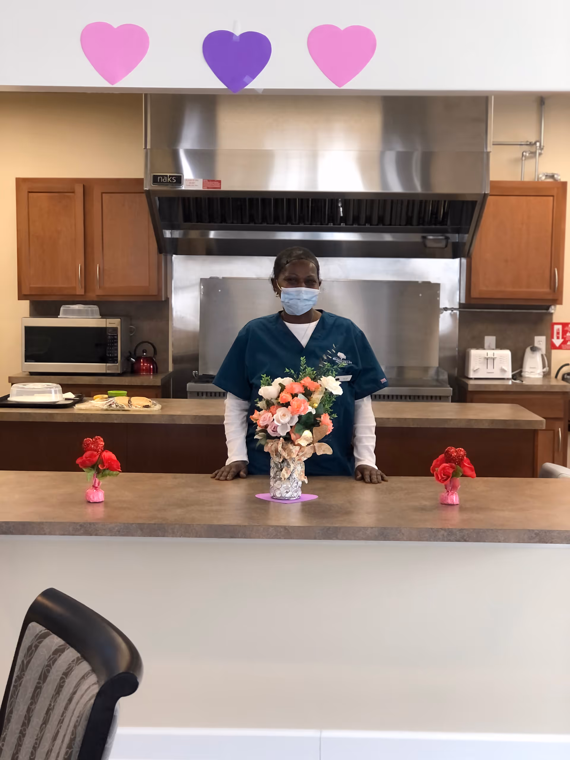 A masked staff member in teal scrubs stands behind a kitchen counter decorated with flower arrangements and paper heart cutouts.