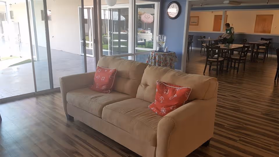 A beige sofa with two red decorative pillows is placed on a wooden floor in a room with large glass sliding doors leading to an outdoor patio. In the background, there is a dining area with several tables and chairs, and a person is seen standing near one of the tables. A small round table with a floral tablecloth and a water dispenser is positioned near the glass doors. A clock is mounted on the blue wall above the table.