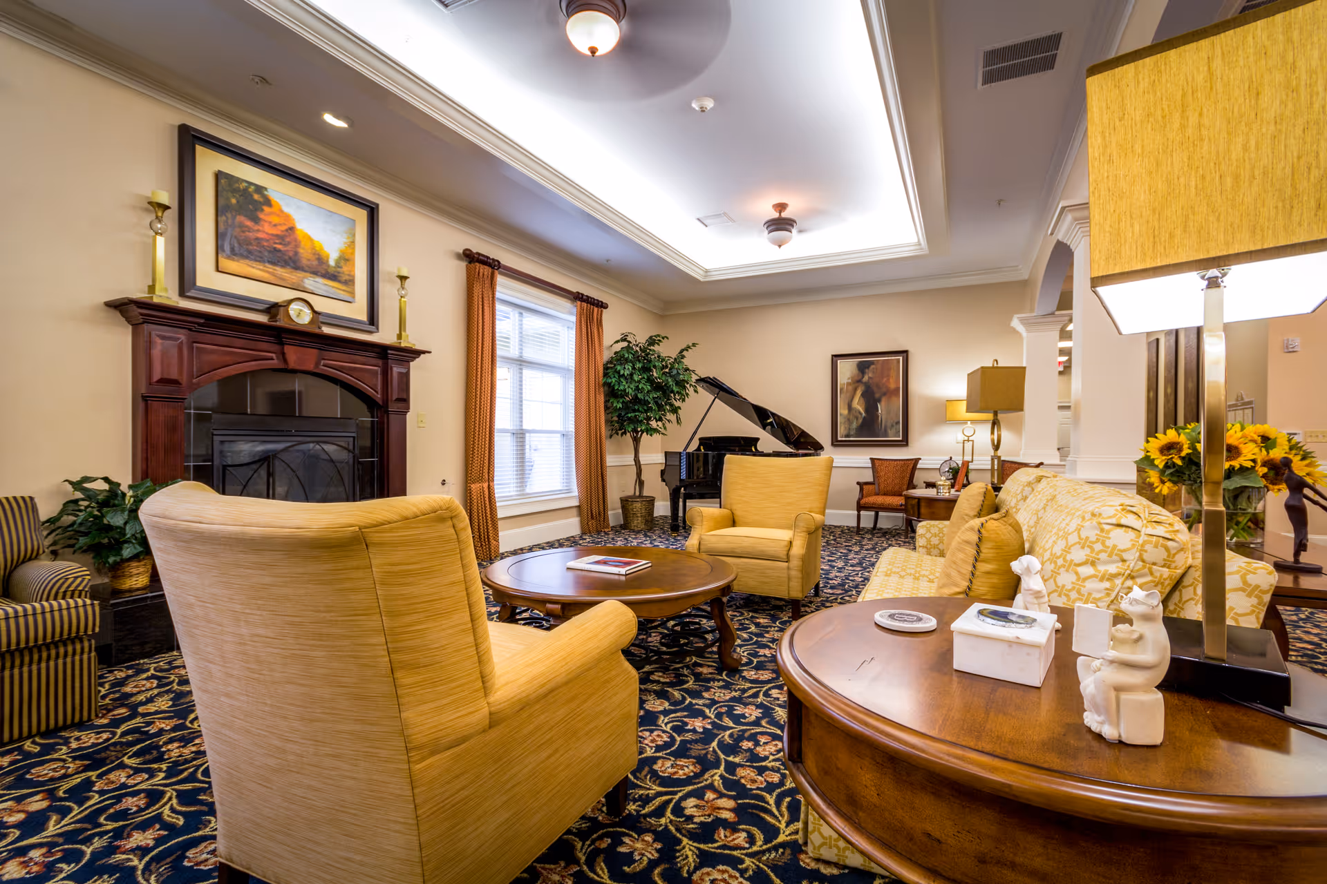 A cozy living room area in a senior living facility featuring yellow upholstered armchairs and a sofa arranged around a wooden coffee table. The room has a dark patterned carpet, a fireplace with a painting above it, large windows with orange curtains, a grand piano in the corner, and decorative lamps and plants.