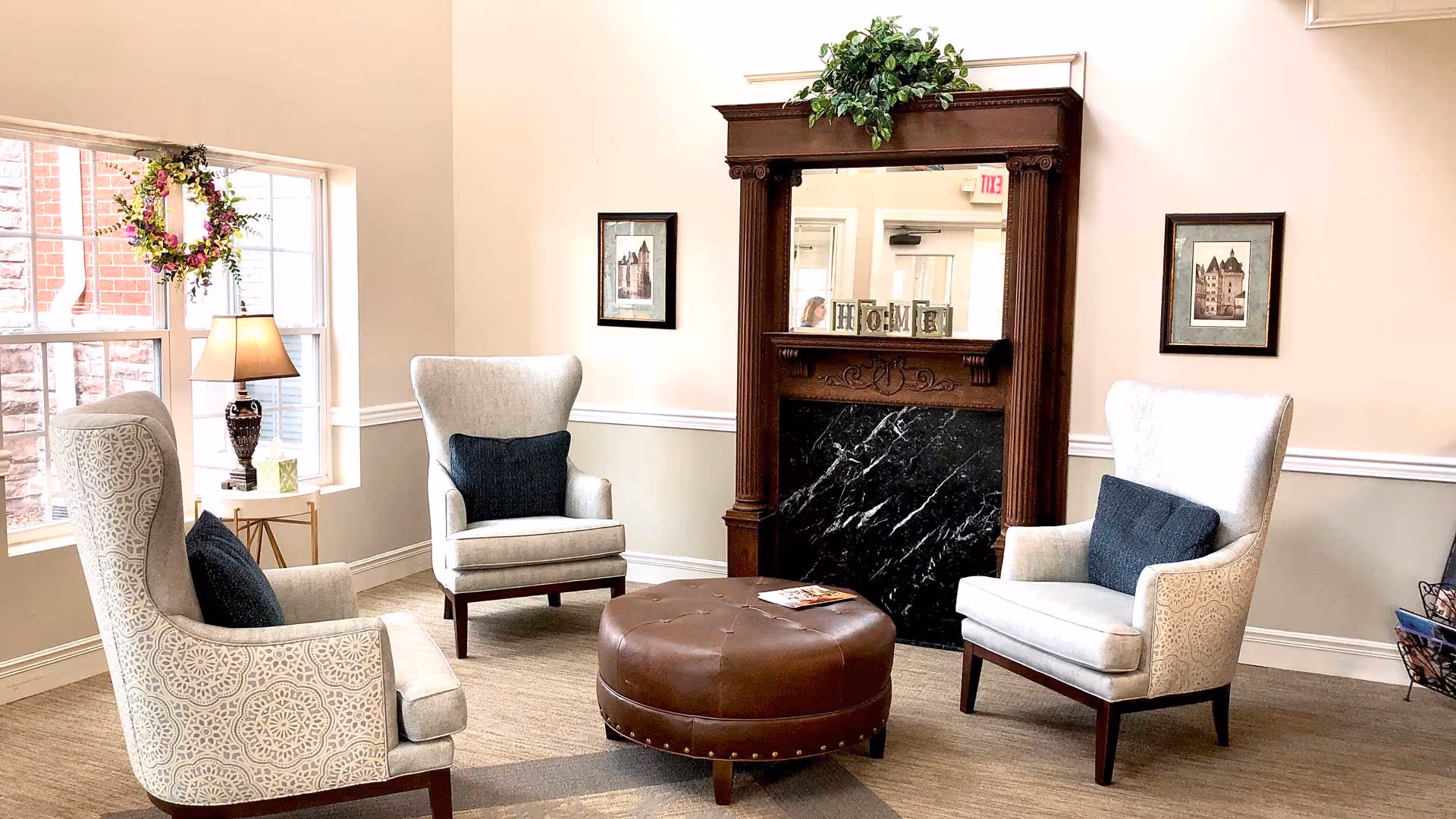 A cozy sitting area with three patterned armchairs arranged around a round brown leather ottoman. Behind the chairs is a decorative wooden fireplace mantel with a black marble insert and a mirror above it. The mantel has decorative blocks spelling 'HOME' and a green plant on top. Two framed pictures hang on the wall on either side of the fireplace. A window to the left lets in natural light, with a floral wreath hanging in front of it and a table lamp on a small side table below.