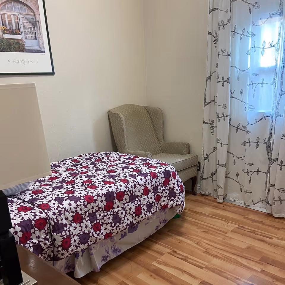 Small bedroom with a single bed covered in a purple and white floral quilt, an upholstered armchair in the corner, and a window with patterned curtains.