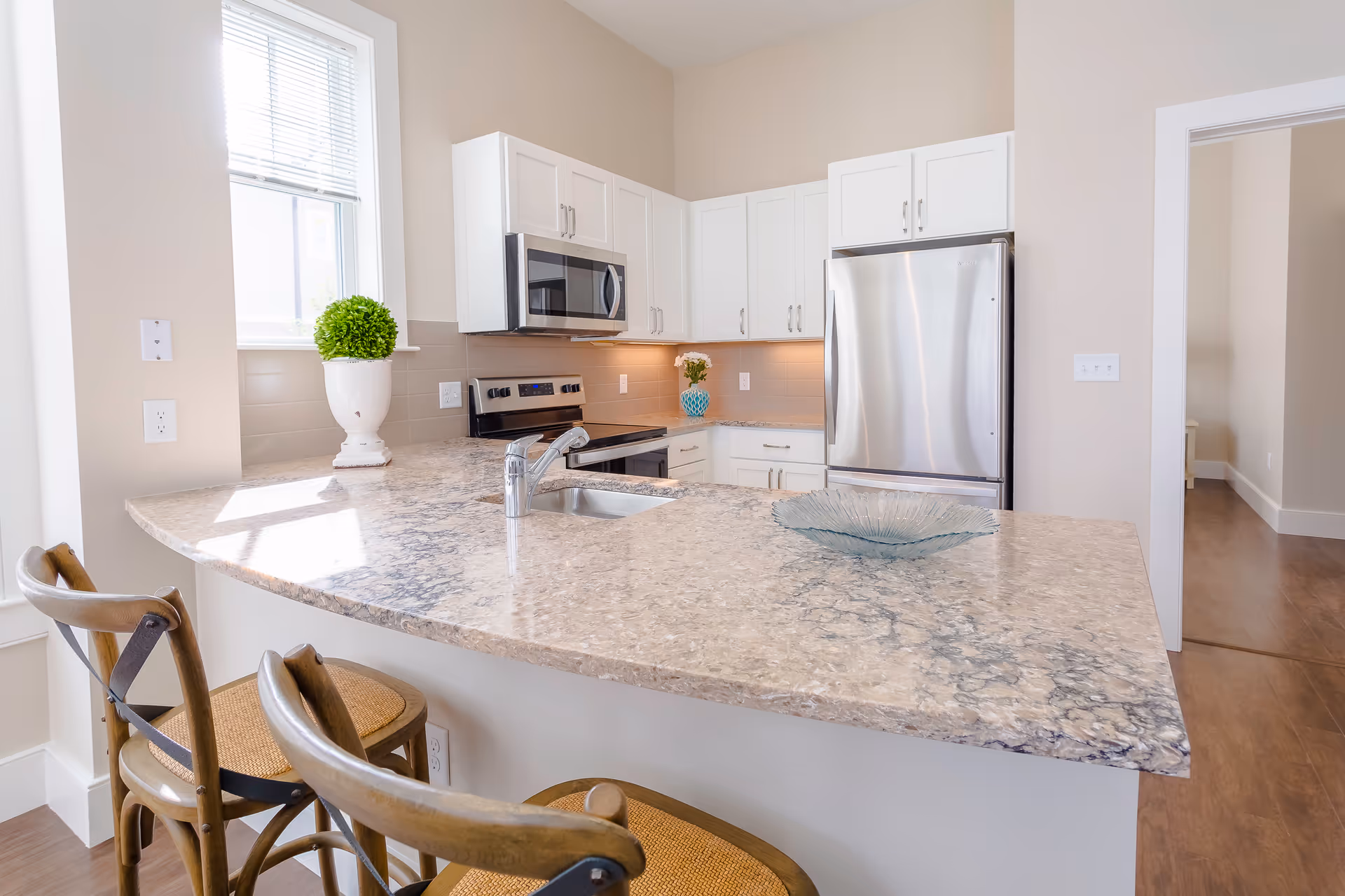 Bright modern kitchen with a marble-topped island, sink, stainless steel appliances, white cabinets, and two bar stools.