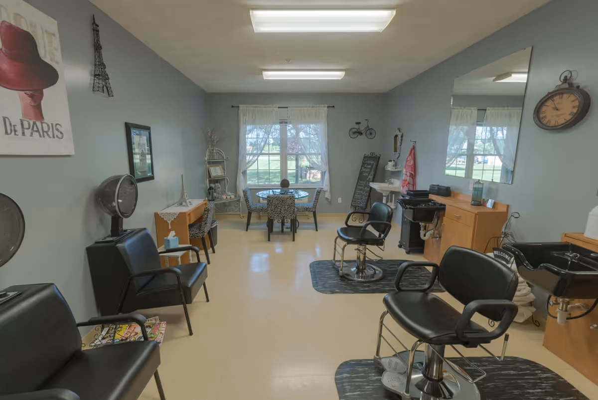 Interior view of a salon area in a senior living facility with two black salon chairs on mats, a large mirror on the wall, a sink for hair washing, and a small table with chairs near a window with sheer curtains. The walls are decorated with Paris-themed artwork and a clock.