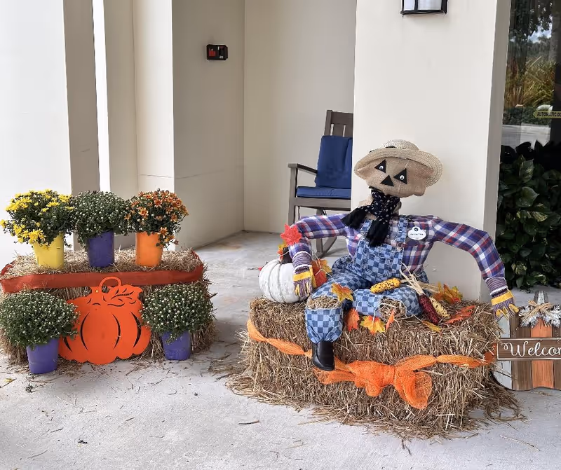 A fall-themed outdoor decoration featuring a scarecrow dressed in blue checkered overalls, a plaid shirt, and a straw hat sitting on a hay bale adorned with autumn leaves and corn. Next to the scarecrow is a display of potted flowers in yellow, purple, and orange pots arranged on hay bales with an orange pumpkin cutout. A wooden chair with a blue cushion is in the background near a white wall.