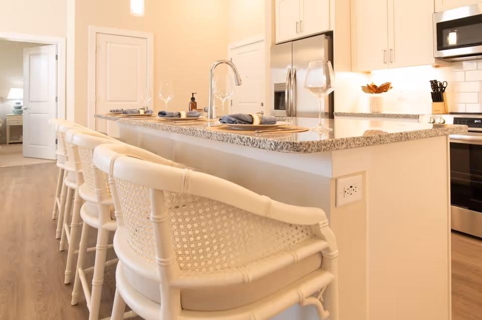 Bright modern kitchen featuring a granite-topped island with four white bar stools, place settings, and stainless steel appliances.