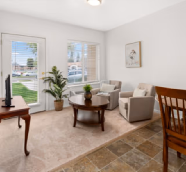 A bright living room with two beige armchairs, a round wooden coffee table with a small plant, a wooden TV stand with a television, a potted plant near a window, and a wooden dining chair partially visible. The room has carpet and tile flooring, white walls, and a window and door with blinds showing an outdoor view.