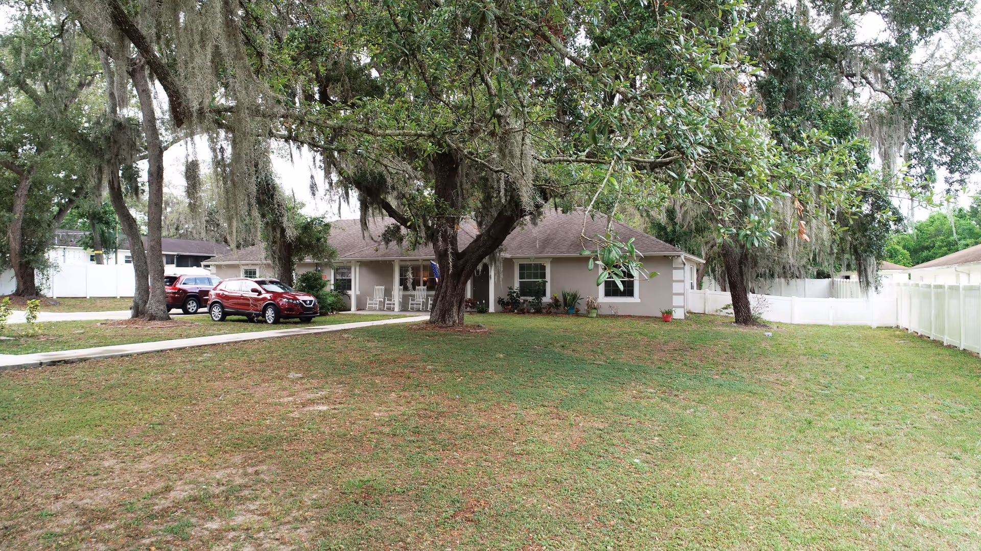 Single-story residential building with a front porch featuring rocking chairs, surrounded by large trees with hanging moss. Two cars are parked on the driveway to the left, and a white fence encloses the property. The lawn is green with some patches of bare ground.