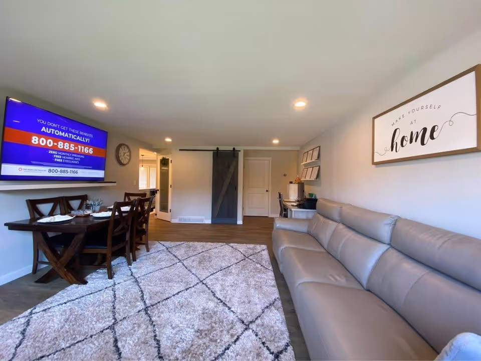 A modern living room with a long gray leather sofa on the right, a large patterned area rug on the floor, and a wooden dining table with chairs on the left. A flat-screen TV is mounted on the wall above the dining table displaying a phone number and advertisement. The back wall features a sliding barn door and a small desk with a chair and computer. A framed sign on the wall reads 'Make yourself at home.'