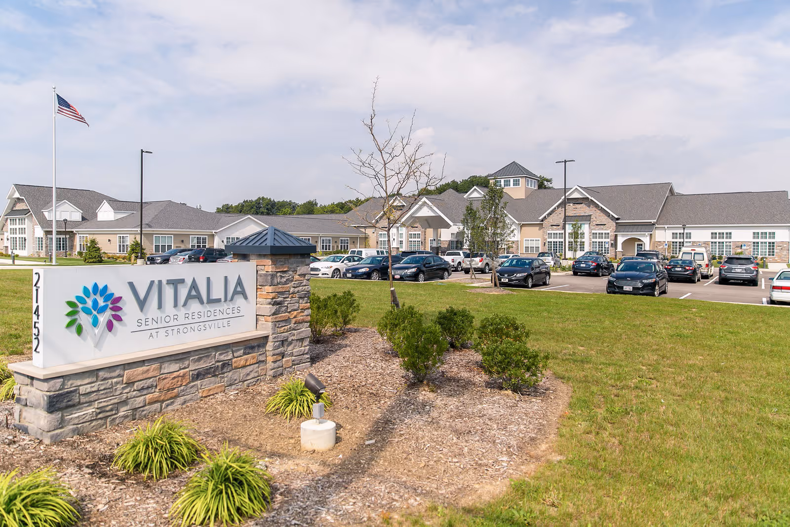 Exterior view of Vitalia Senior Residences at Strongsville showing a large building with multiple windows, a parking lot with several cars, a landscaped area with small bushes and grass, and a sign with the facility's name and logo. An American flag is flying on a flagpole near the sign.