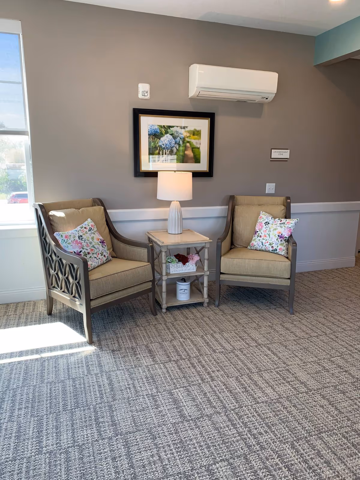 Two upholstered armchairs with floral pillows flank a small side table and lamp beneath framed artwork in a bright sitting area.