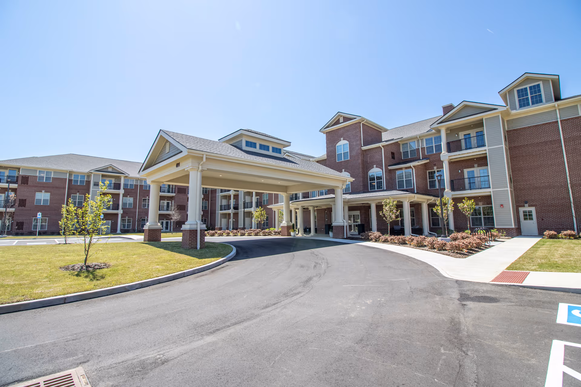 Exterior view of a large senior living facility building with a covered entrance driveway, multiple windows, and balconies. The building is surrounded by a well-maintained lawn, small trees, and a clear blue sky.