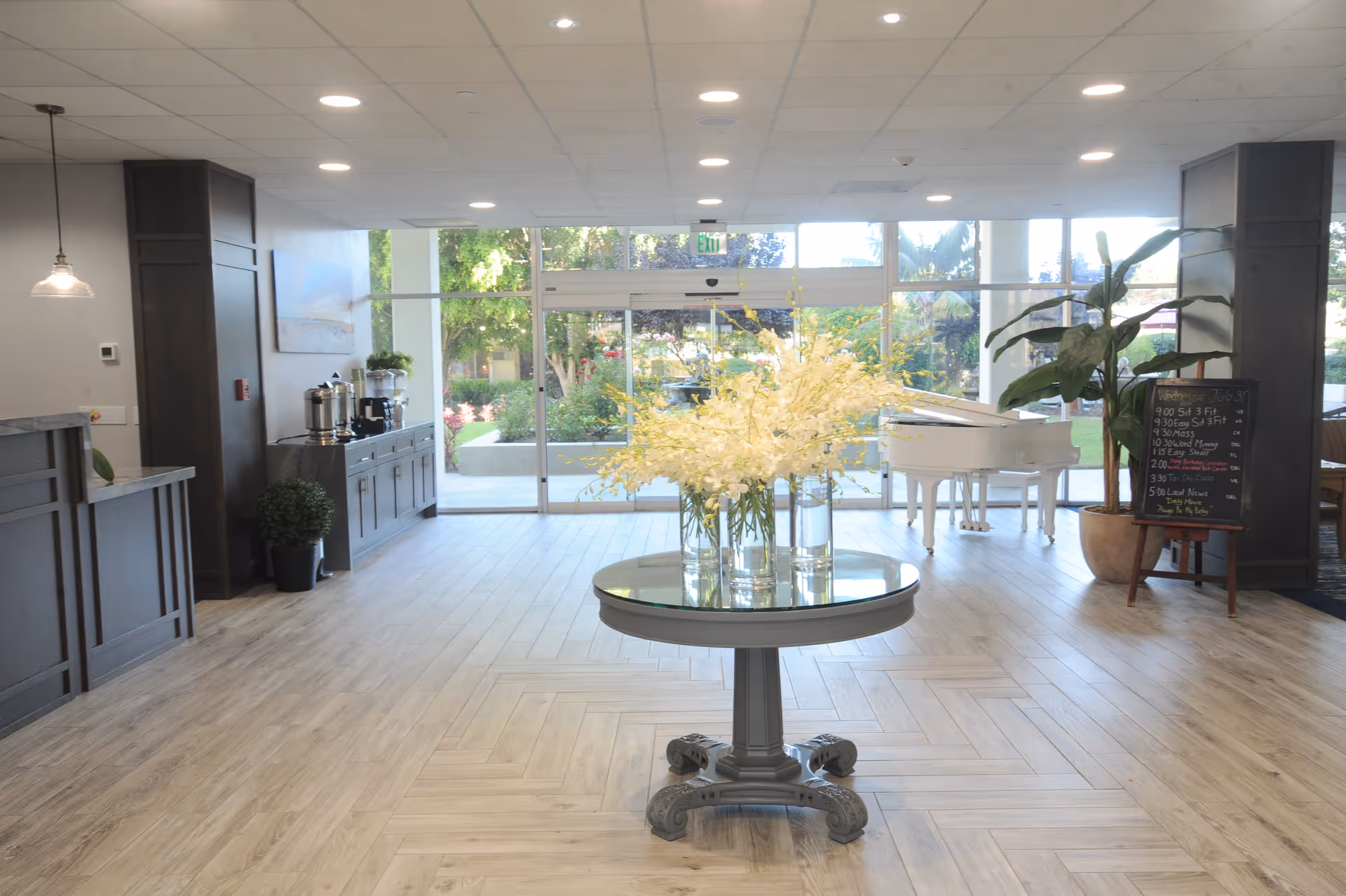Bright and spacious lobby area with light wood flooring, a round table with vases of white flowers in the center, a white grand piano near large glass doors leading outside, a chalkboard with daily activities, and a sideboard with coffee and tea setup.
