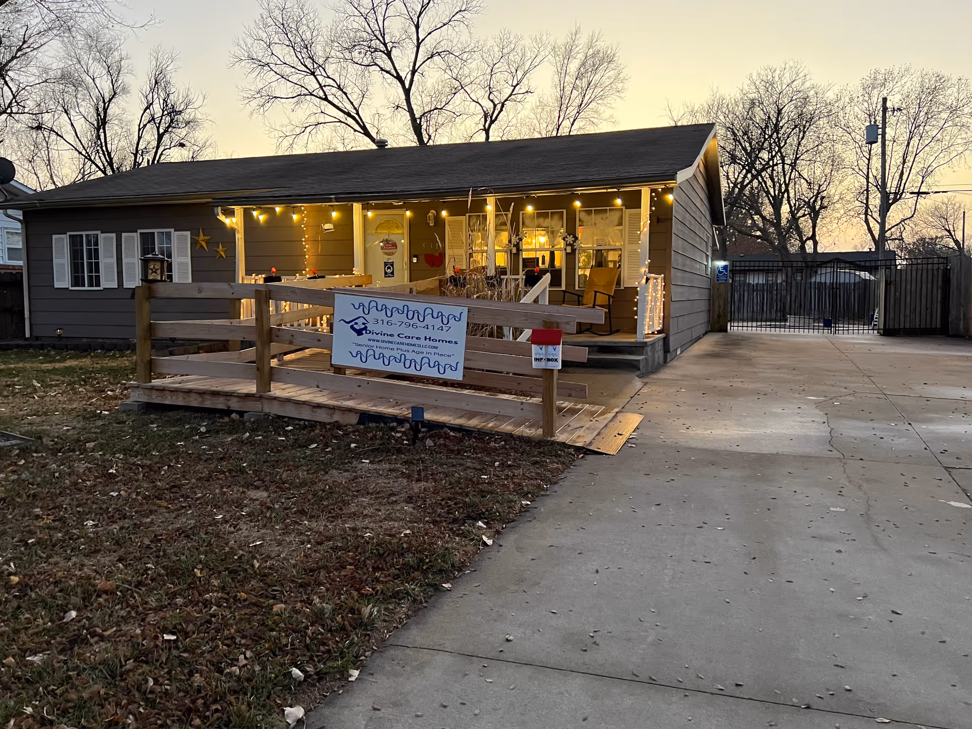 Exterior view of a single-story house at dusk with a wooden ramp leading to the front porch. The porch is decorated with string lights and has a rocking chair. There is a sign on the ramp that reads 'Divine Care Homes' with a phone number. The driveway is concrete and leads to a gated area. Leafless trees are visible in the background.