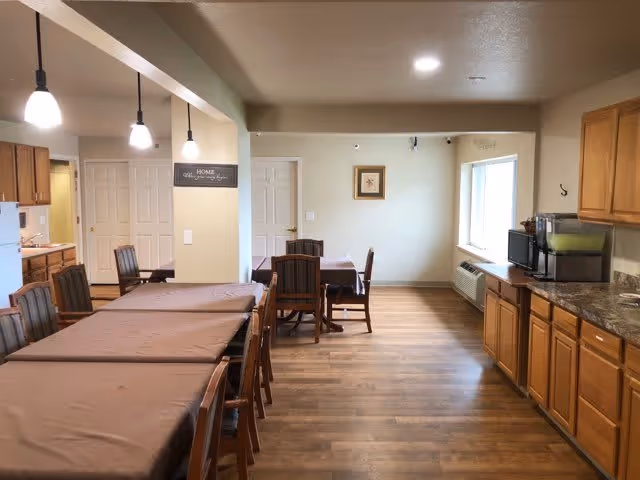 Communal dining area with multiple covered tables and chairs, pendant lights, and a kitchenette with wooden cabinets along the right wall.