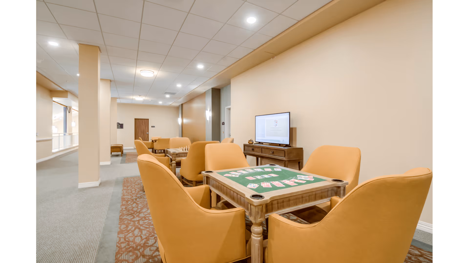 A well-lit common area with beige walls and ceiling lights, featuring a card table with playing cards and four orange upholstered chairs around it. In the background, there are additional seating areas with similar chairs and tables, and a TV mounted on a wooden console against the wall.