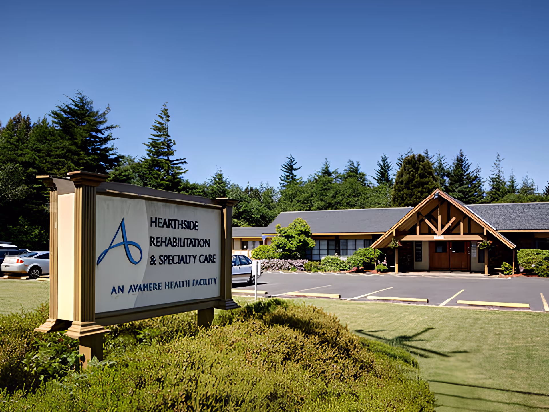 Exterior view of Hearthside Rehabilitation & Specialty Care, an Avamere Health Facility, showing the building entrance with a peaked wooden awning, surrounded by greenery and a parking lot with several cars under a clear blue sky.