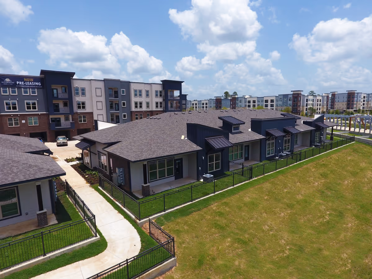 Aerial view of a senior living facility named Watermere at Woodland Lakes showing multiple residential buildings with fenced yards, a paved walkway, and a grassy area under a partly cloudy sky.
