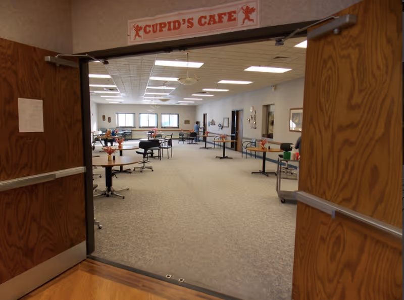 View through double doors into a large community dining/cafe room labeled 'Cupid's Cafe' with scattered tables and chairs.