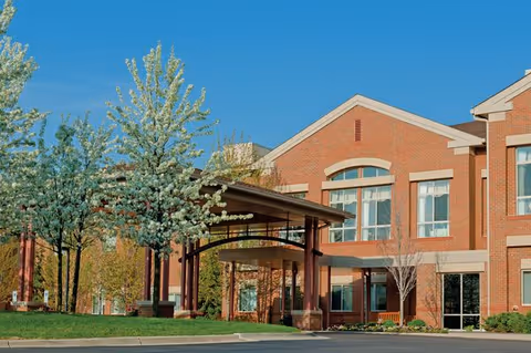 Exterior view of a brick senior living community building with large windows and a covered entrance. Trees with white blossoms and green grass are visible in the foreground under a clear blue sky.