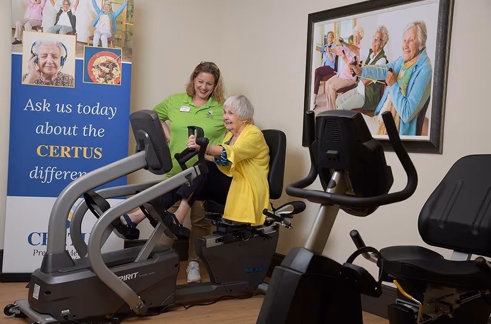 An elderly woman in a yellow cardigan is using a recumbent exercise bike in a fitness room, assisted by a smiling staff member in a green shirt. Behind them is a blue banner with text about CERTUS and a framed photo of elderly people exercising with resistance bands on the wall.